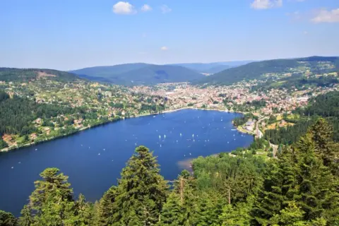 Vue panoramique du lac de Gérardmer, avec des voiliers sur l'eau bleue, entouré de collines verdoyantes et d'une ville pittoresque.