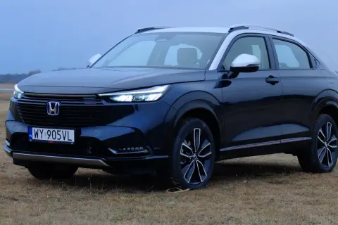 Nowa Honda HR-V w ciemnym kolorze, zaparkowana na trawiastym polu, z widokiem na jezioro i las w tle.