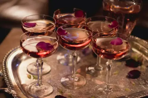 Eleganckie kieliszki z różowym winem, ozdobione płatkami róży, czekają na degustację. W tle widać karafkę z tym samym trunkiem, być może to wykwintny tokaji aszu.