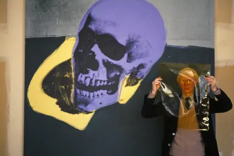 Andy Warhol trzyma przezroczystą folię przed fioletowo-żółtym obrazem czaszki.