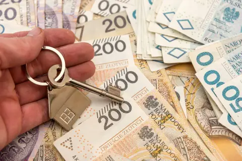 Dłoń trzyma klucz z breloczkiem w kształcie domu na tle polskich banknotów. Symbolizuje to możliwość zakupu nieruchomości lub dziedziczenia, co wiąże się z kwestią zachowku.