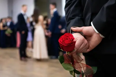 Elegancki mężczyzna w garniturze trzyma za plecami czerwoną różę, symboliczny gest na dzień mężczyzny w marcu. W tle widać bawiących się ludzi.
