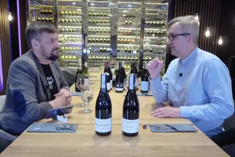 Dwóch mężczyzn przy stole degustuje wino. Na stole stoją butelki, w tym dwie z etykietami sugerującymi pinot noir.