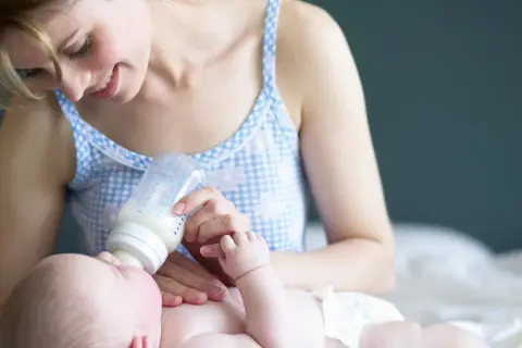 Uśmiechnięta mama karmi butelką noworodka. Warto wiedzieć, ile powinien jeść dwutygodniowy noworodek, by zapewnić mu prawidłowy rozwój.