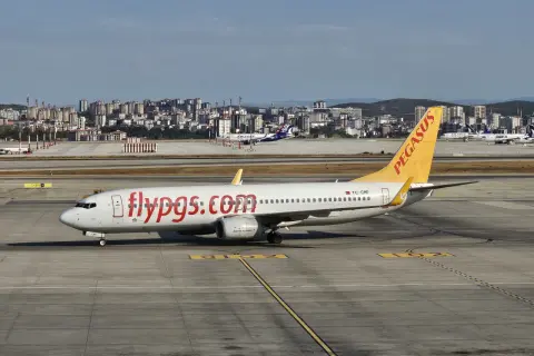 Samolot linii lotniczych Pegasus z napisem "flypgs.com" na kadłubie na płycie lotniska, w tle miasto.