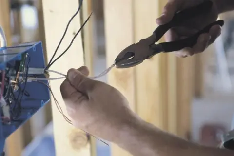 Dłonie trzymające kombinerki i przewody elektryczne przy niebieskiej puszce instalacyjnej.