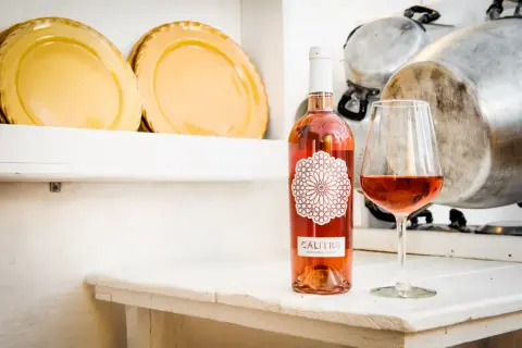 Un bicchiere e una bottiglia di negroamaro rosato Calitro su un tavolo bianco, con piatti gialli e pentole sullo sfondo.