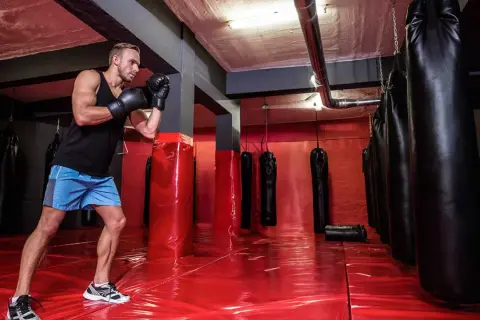 A boxer trains in a gym with many boxing bags. The red floor reflects the lights, and the boxing bag numbers are not visible.
