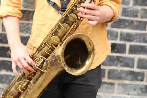 Ein Musiker spielt ein goldenes Saxophon vor einer Ziegelwand. Hier ist ein Saxophon Steckbrief: ein Blasinstrument mit Klappen und einem Mundstück.