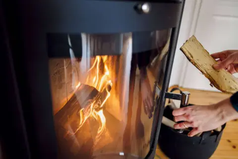 Eine Hand legt Holzscheite in einen brennenden Ofen. Die Flammen lodern im Inneren des Ofens.