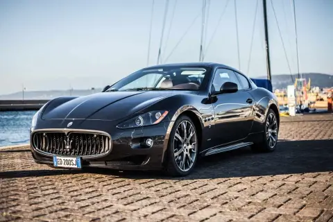 Elegancki, czarny Maserati coupe lśni w słońcu na tle portu z żaglówkami.