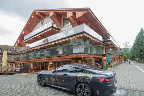 Luksusowy hotel Górski Diament z nowoczesną architekturą i przeszklonymi balkonami. Przed budynkiem stoi elegancki samochód z motywem "Księstwo Góralskie".