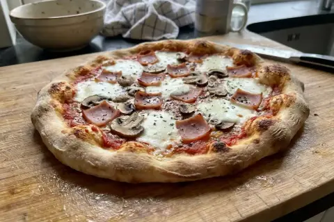 En nybakad pizza med tomatsås, mozzarella, skinka och champinjoner ligger på en skärbräda.