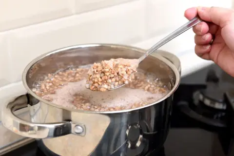 Eine Hand hebt einen Löffel mit dampfendem Buchweizen aus einem Topf, in dem Buchweizen gekocht wird.