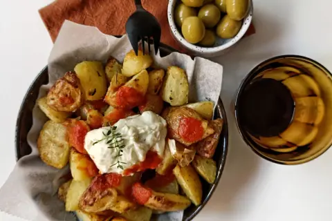 Patatas bravas con alioli, servidas con aceitunas y una bebida oscura.