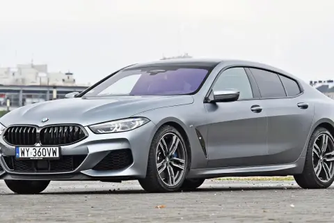 Szary BMW M8 Gran Coupe z czarnym dachem i felgami, zaparkowany na ulicy.