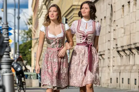 Zwei Frauen in Dirndln ohne Schnürung spazieren. Die Kleider sind in Rosa- und Beigetönen gehalten und mit zarten Stickereien verziert.