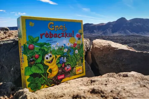Gnaj, robaczku!" - planszówka z wesołymi robaczkami i truskawkami, na tle surowego krajobrazu.