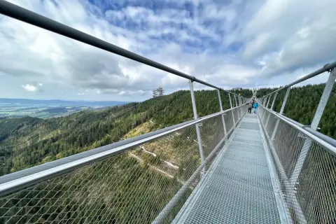 Himmelbrücke in Dolní Morava mit Blick auf bewaldete Hügel und eine Fernsehturm-ähnliche Struktur.