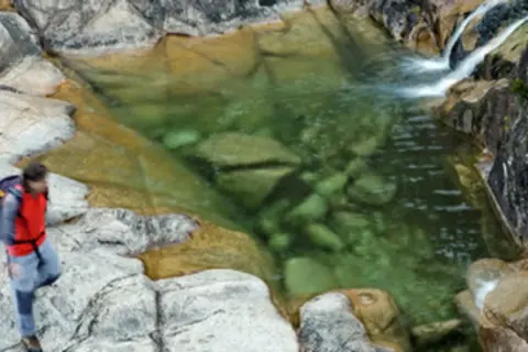 Senderista se detiene junto a un **río caldo** de aguas cristalinas y rocas musgosas.