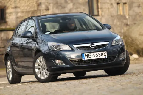 Ciemny Opel Astra J z włączonymi światłami do jazdy dziennej, zaparkowany na kamienistej drodze.