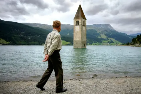 Ein Mann spaziert am Ufer eines Sees, auf dem ein Kirchturm aus dem Wasser ragt. Die Kirche im Stausee ist ein eindrucksvolles Zeugnis der Vergangenheit.