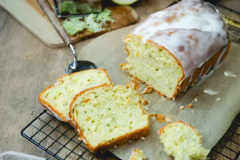 Saftiger Zucchini Kuchen mit Zuckerguss, frisch angeschnitten auf einem Gitter. Daneben liegen geriebene Zucchini und ein Hobel.