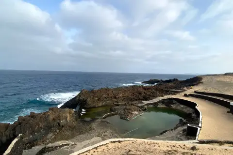 Piscinas naturales formadas por lava volcánica en la costa de Lanzarote, con el mar azul de fondo. No hay cocoteros, pero sí un paisaje salvaje.