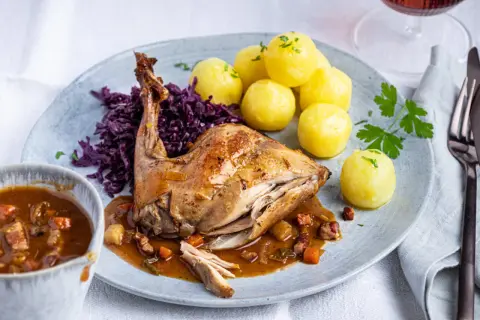 Rezept Hasenbraten im Backofen: Saftiges Kaninchen mit Klößen, Rotkohl und Sauce auf einem Teller.