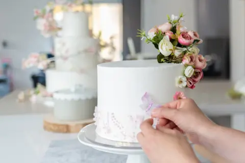 Una mano aggiunge una farfalla a una torta bianca decorata con fiori e paillettes. Sullo sfondo, un'altra torta a più piani con decorazioni floreali rosa.