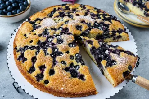 Saftiger Blaubeerkuchen, bestäubt mit Puderzucker. Ein Stück wird gerade herausgehoben, daneben eine Schüssel voller frischer Blaubeeren.