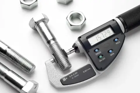 Cyfrowy mikrometr mierzy śrubę z gwintem m12, pokazując 15.729 mm. Wokół leżą nakrętki.