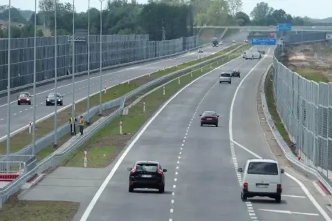 Samochody poruszają się lewym pasem w terenie zabudowanym, mijając ekrany akustyczne.