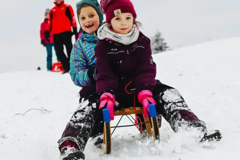 Zwei Kinder rodeln lachend einen verschneiten Hang im Bergischen Land hinunter.