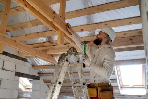 Obrero con casco y martillo realiza refuerzo estructural de vigas de madera en una construcción.