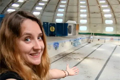 Une jeune femme souriante explore une piscine tournesol abandonnée, son dôme blanc orné de fenêtres rondes.