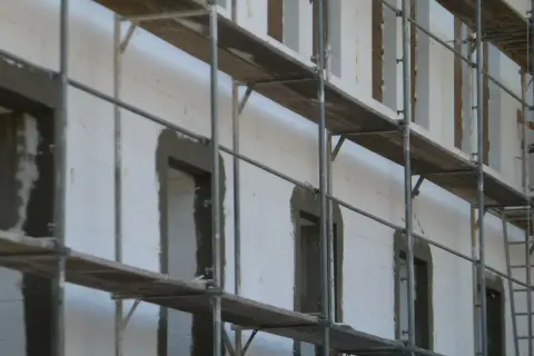 Budowa domu z bloczków styropianowych. Rusztowania otaczają białą ścianę z otworami okiennymi i drzwiami, przygotowanymi pod dalsze prace.