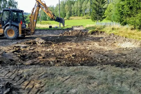 Koparka pracuje, tworząc wykopy pod fundamenty. Teren jest błotnisty, z gruzem i kamieniami.