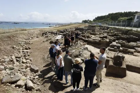 Grupo de personas observando ruinas arqueológicas en la costa. Se aprecia el mar y el **adro vello** del yacimiento.