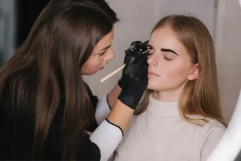 Profesjonalne farbowanie brwi. Stylistka w czarnych rękawiczkach precyzyjnie maluje brwi klientce.