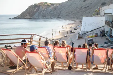 Gente relajándose en tumbonas con vistas a la playa de Burgau. El mar azul y la costa rocosa crean un paisaje idílico.