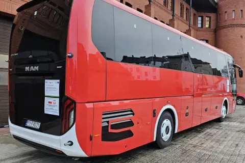 Czerwony autokar MAN Lion's Coach zaparkowany przed ceglanym budynkiem. Może to być przystanek w podróży z Bolesławca do Poznania.