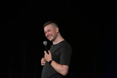 Grzegorz Wójtowicz w trakcie występu stand-up, trzyma mikrofon i uśmiecha się do publiczności.