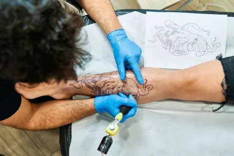 Tätowierer mit blauen Handschuhen sticht Oktopus-Motiv auf Bein. Tolle Tattoo-Ideen für Meeresliebhaber.