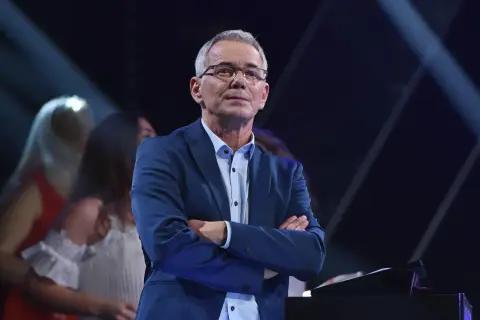 Robert Janowski w garniturze, z rękami skrzyżowanymi na piersi, na tle sceny. Czy to właśnie powód, dlaczego Robert Janowski naprawdę odszedł z Jaka to melodia po latach?