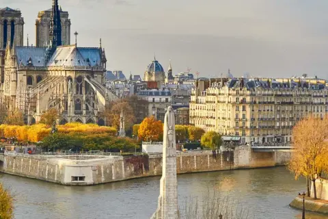 Vue panoramique de Paris en automne, avec la majestueuse Notre Dame de Paris dominant le paysage, bordée par la Seine et des arbres aux couleurs flamboyantes.