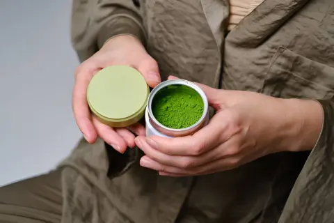 Mains tenant une boîte ouverte de poudre de matcha verte vibrante, évoquant les matcha bienfaits peau.