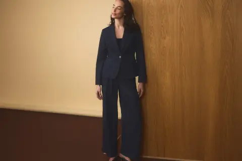 Elegancka kobieta w granatowym garniturze, z kobaltową marynarką damską i szerokimi spodniami, opiera się o drewnianą ścianę.