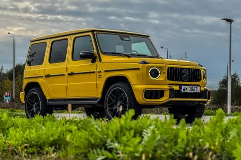 Żółty Mercedes-Benz G63 AMG z czarnymi felgami i przyciemnianymi szybami, zaparkowany na tle pochmurnego nieba i zieleni.