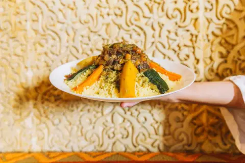 Un plat de couscous algérien, garni de légumes colorés et d'une viande savoureuse, est présenté sur un fond orné.
